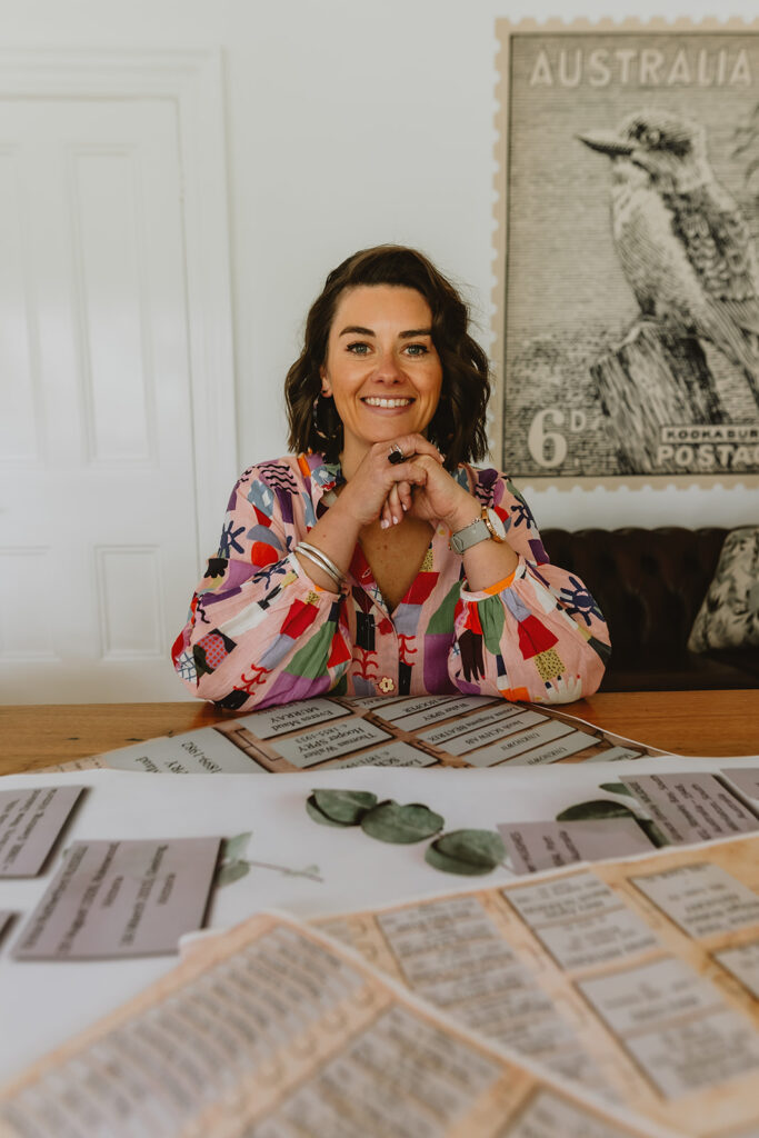 Phoebe sits at a wooden table with different family trees muted in front of her in different artwork options.  Wearing a brightly coloured dress with a white backdrop of the walls you can also see a brown chesterfield couch and a big stamp on the wall with a kookaburra on it.