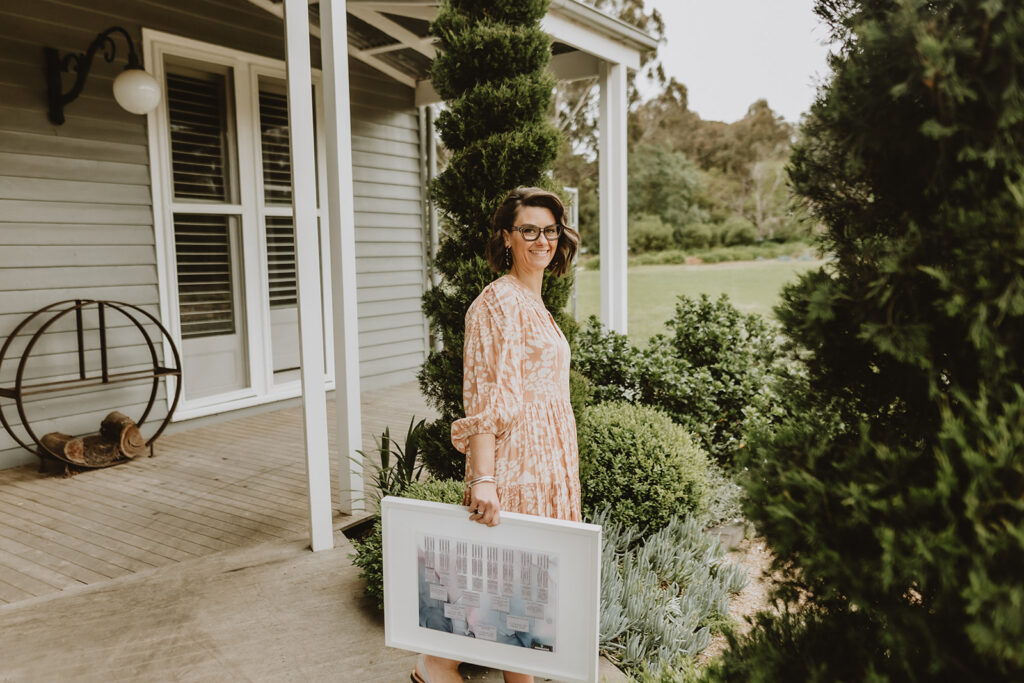 Phoebe standing on a verandah holding a white large sized frame with a family tree inside of it, wearing a long flowy muted orange dress with a white print on it.  In the background you can see green grass, and a tree in the foreground at the front of a muted grey weatherboard house.