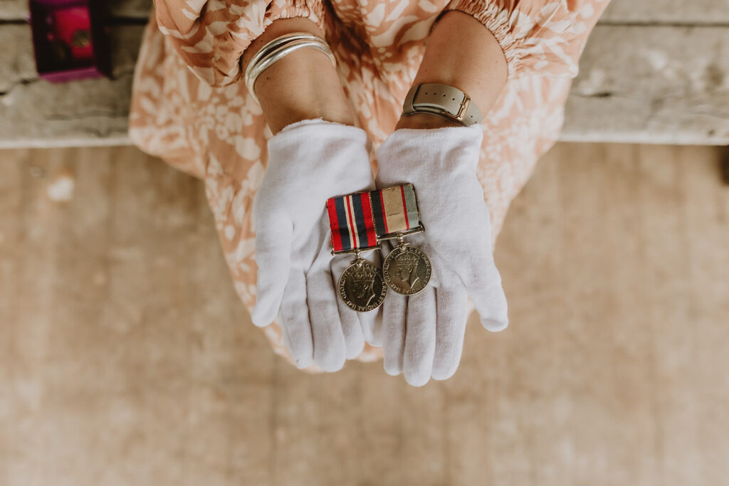 Phoebe holding two war medals in white protective gloves whilst sitting on a bench seat along a verandah.  Phoebe is wearing a muted orange tone dress with a lovely white floral print, you can see two silver bangles on Phoebe's wrist and her watch on the other.
