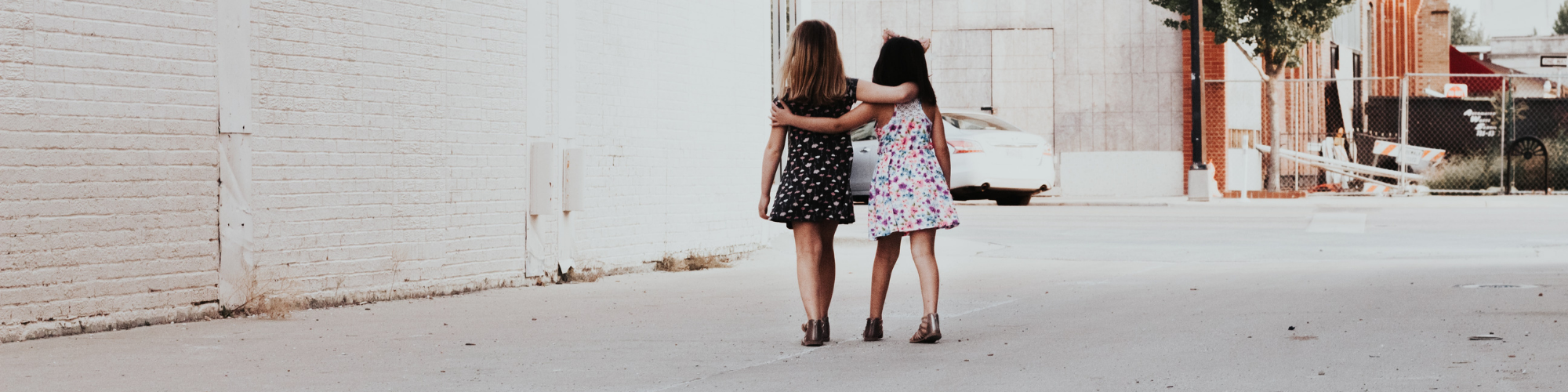 Pictured two little girls, both wearing dresses and flat shoes walking away from the camera with their arms over each others shoulders