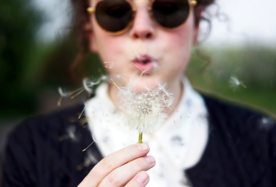 Woman out of focus wearing a black jumper with a white coloured shirt underneath, wearing tortoise shell coloured sunglasses, blowing a dandelion and making a wish