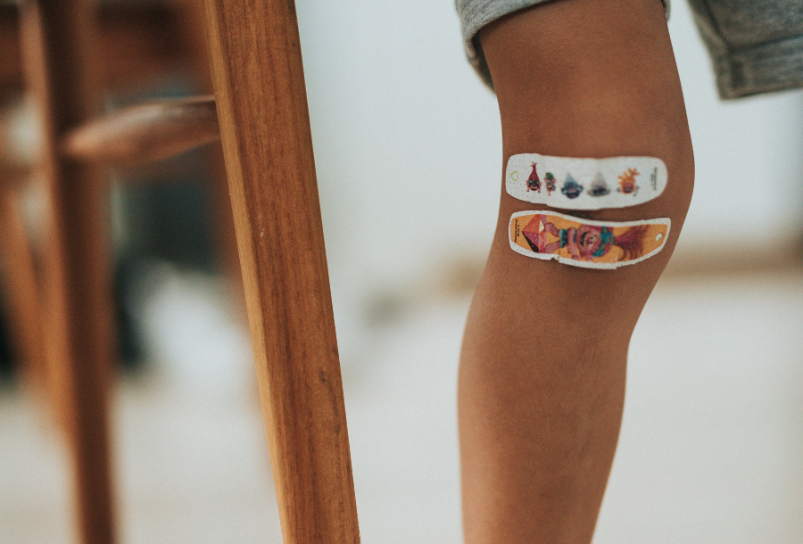 Close up picture of a young child's leg sitting on a wooden chair with two bandaids on the bend of the knee, the top one is white with a small print and the bottom one is orange with a white outline