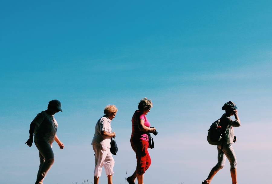 Picture of four retirement aged women who are spread out across a beautiful blue sky with backpacks on, enjoying a lovely afternoon walk in the sunshine