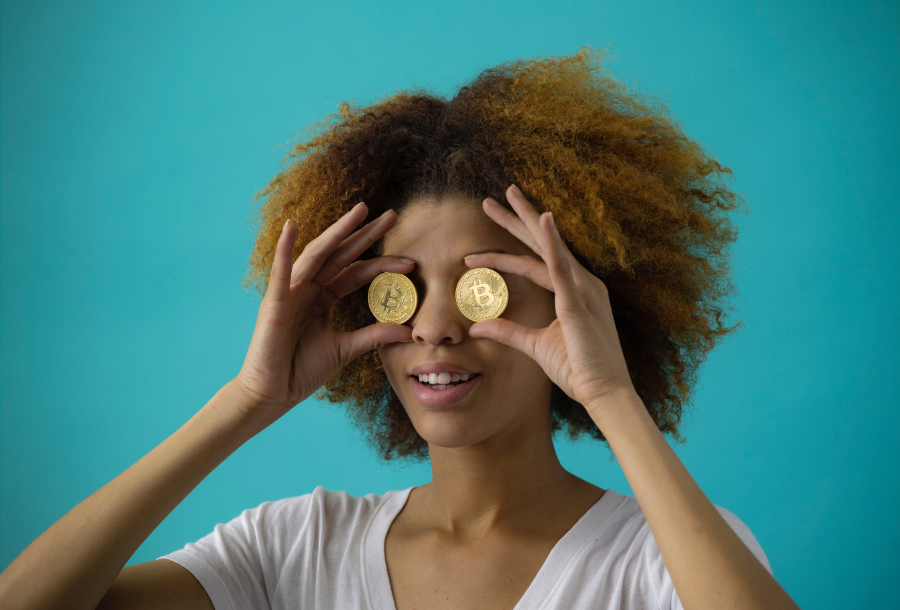 Picture of a girl who is holding up gold "bitcoins" up to her eyes, she has amazing curly big brown hair and is wearing a white round low neck t-shirt