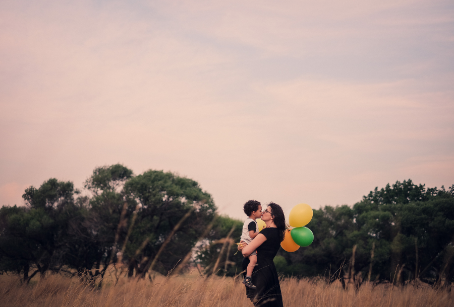 Picture of a woman holding a young child in her arms and the child is holding a bunch of orange, yellow and green balloons while they standing in a field at sunset with beautiful green trees in the background