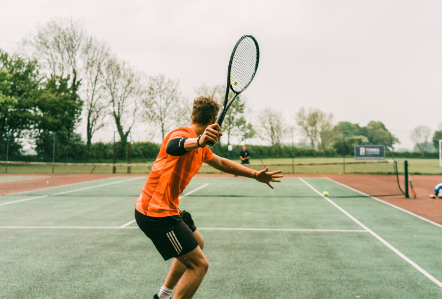 Picture of a green grass tennis court with orange trimmings, a person wearing an orange top and black sports pants holding a tennis racket in his right hand about to hit the ball