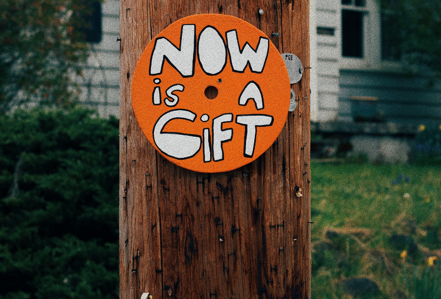 Orange coloured round circle stuck to a power pole with the words "now is a gift" in white writting and black outlined, a white weatherboard house in the background with green grass