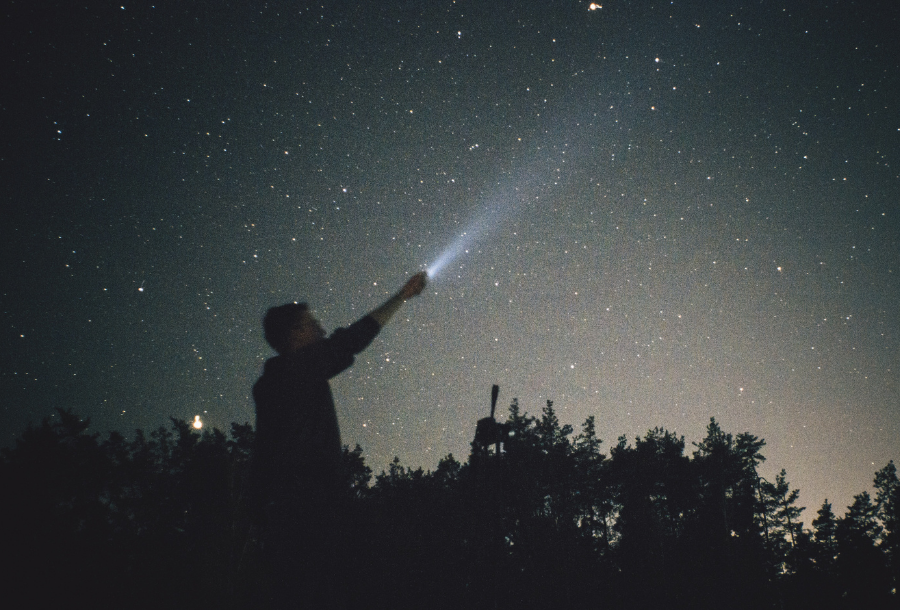 Picture of a person standing outside in the dark night sky holding up a torch into the sky to point towards the stars which are filling the night sky.