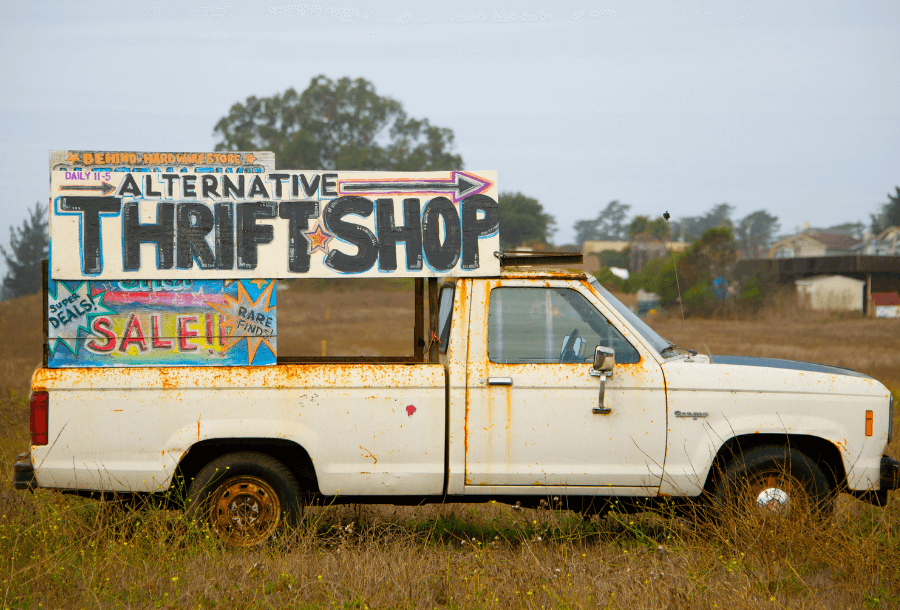 White ute sitting in a field next to the road, with rust running down the side of the tray and from the door hands, with a sign on the roof that says "Alternative Thrift Shop" with an arrow point right and a sign underneath with bright starts that says "sale"