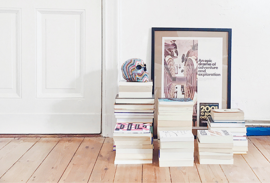 IMAGE DESCRIPTION: five piles of books stacked at different heights on beautifully polished floorboards with a piece of artwork leaning up against a white wall and a rainbow colour skull head on the highest pile of books