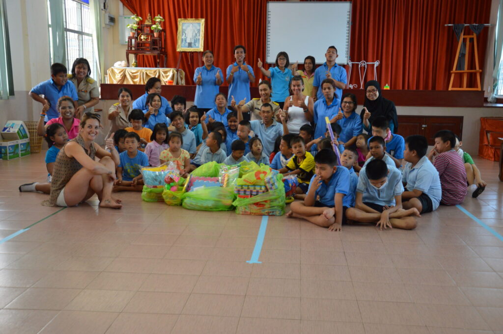 Phoebe Wilkens and Donna Lindsay surrounded by approximately 30 children wearing light royal blue polo tops sitting on the floor in a gym in Thailand, in the background you can see big red heavy curtains with donations at the front of the group