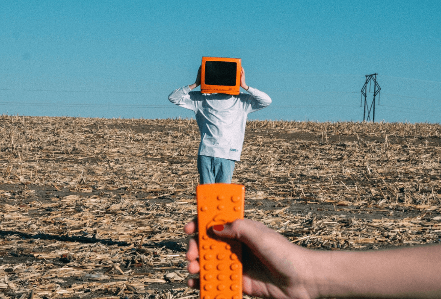 A field that has been harvested, with a beautiful blue sky backdrop and you can see a powerline in the back right hand side of the image, with a person in the centre of the frame holding an orange television covering their face. In the foreground there is an arm holding a matching orange remote control