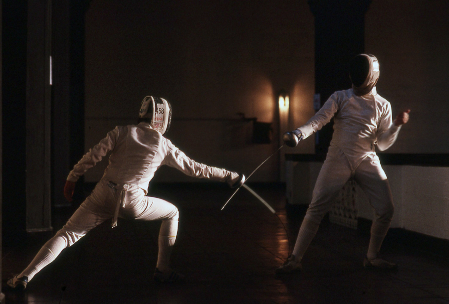 Two fencing people facing off against each other in full while uniform against a dark coloured background, person in the forefront is taking a strong lead on their front right foot and a low stand. A dull light can be seen between the paid against the dark wall in the background