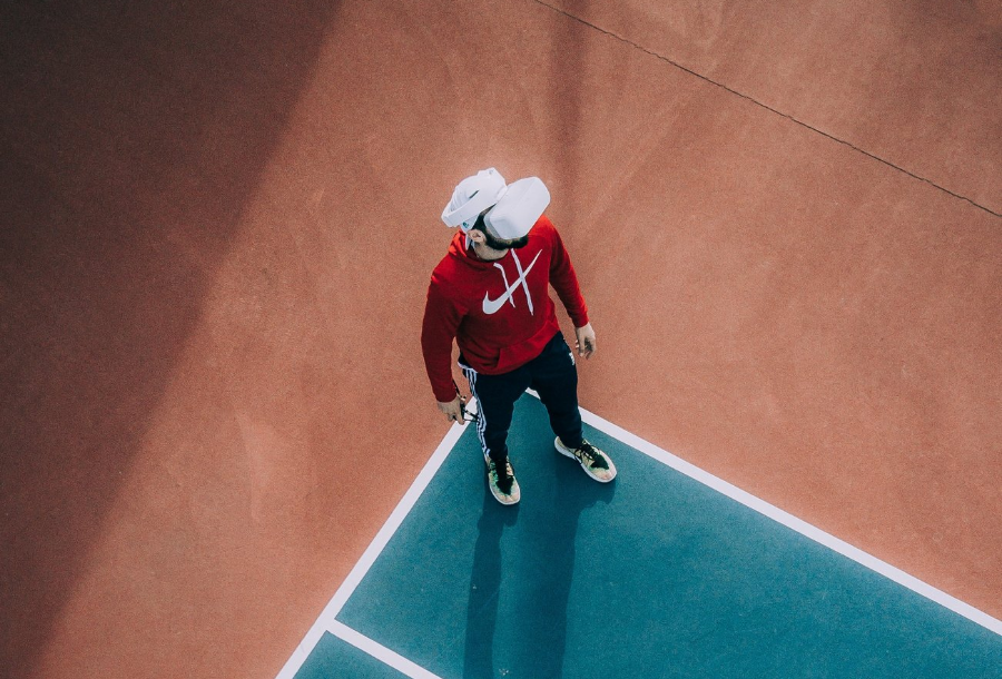 Man wearing a bright red Nike hoodie with white timings, a white baseball cap backwards with black tracksuit pants on standing in the corner of a tennis court with a rust colours outside edge wearing virtual reality goggles