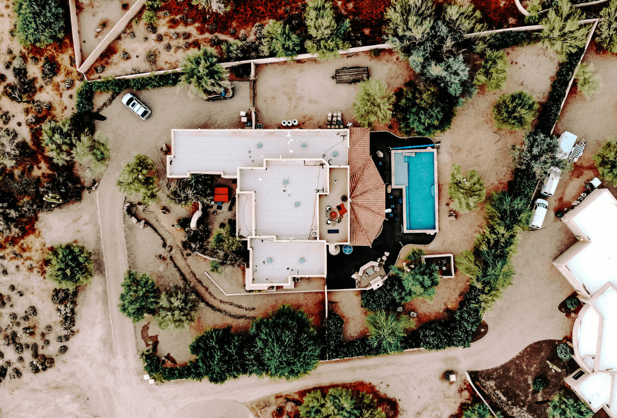 Ariel view of a property, white roof with three large covered areas and an open alfresco area with a pool to the right hand side, beautiful colours of blue. Open landscape around the house with some greenery vegetation around the property