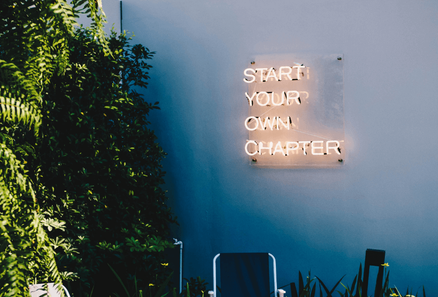 Blank concrete wall with a yellow coloured neon sign that says "START YOUR OWN CHAPTER" lit up with beautiful greenery on the left hand side of the image with the sun reflecting onto it and a couple of comfy camping chairs in the background centre