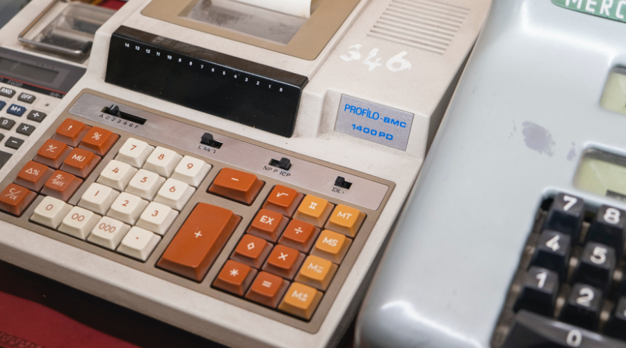 Picture of three calculators that are from an old era, a small one on the left, a big adding machine with orange, white and yellow buttons in the middle and a silver adding machine with black numbers on the right