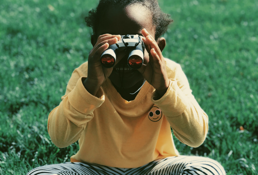 Picture of a young girl looking directly at the camera holding a pair of binoculars, wearing a bright yellow long sleeve top with a white smiley face on the top right hand side near her shoulder and black and white leggings