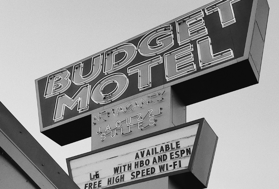 Black and white photo taken from the ground look up towards a big "budget motel" sign with a no vacancy sign and below on a white background, unreadable as there are missing letters