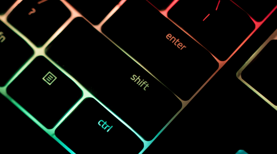 Black keyboard keys that are lit up with neon colours behind the keys, focused on the enter and shift yey with bright blue, green, red and orange colours coming through from behind the black keys