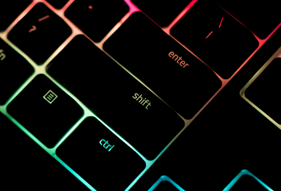 Black keyboard keys that are lit up with neon colours behind the keys, focused on the enter and shift yey with bright blue, green, red and orange colours coming through from behind the black keys