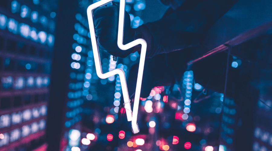White neon lightning bolt in the foreground with a busy background of city lights and car break lights in the bottom of the background of the picture