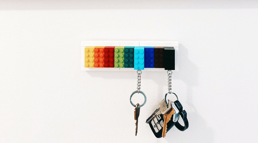 Two sets of keys hanging on a key rings placed against a white washed wall, the keys are hanging from a key holder made of yellow, orange, red, shades of green and blue lego pieces