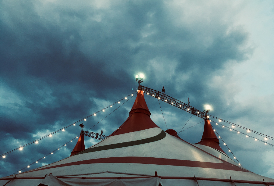 A big reg, white and blue stripped circus tent surrounded by platoon lights against a moody deep blue sky with white clouds