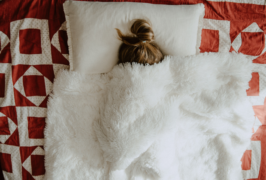 Picture of a person laying on a bed under a white fluffy blanket with only their ponytail showing against a white pillow and a red coloured doona underneath