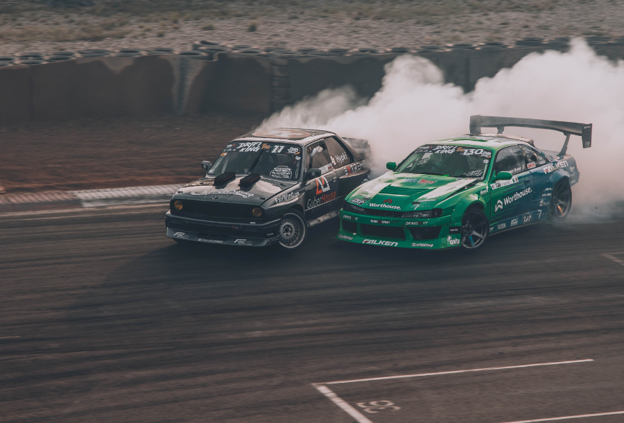 Two racing cars pictured almost side by side, one black with coloured writing over it, the other hot green both drifting around a corner with smoke coming from their tyres