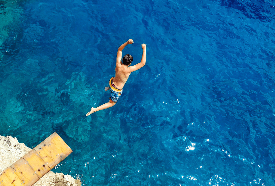 Picture of a young boy jumping off a stone coloured diving board over rocks into the beautiful blue ocean wearing blue and red striped boardshorts