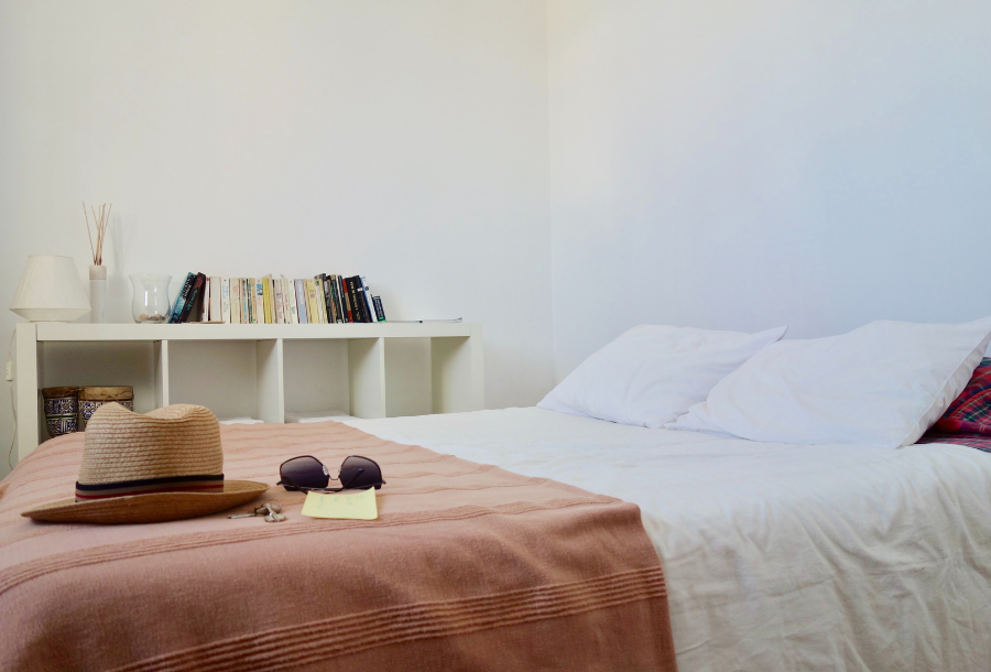 Pictured, a bed made with white sheets and a blush pink throw at the foot with a hat, sunglasses and a note with a bookshelf in the background