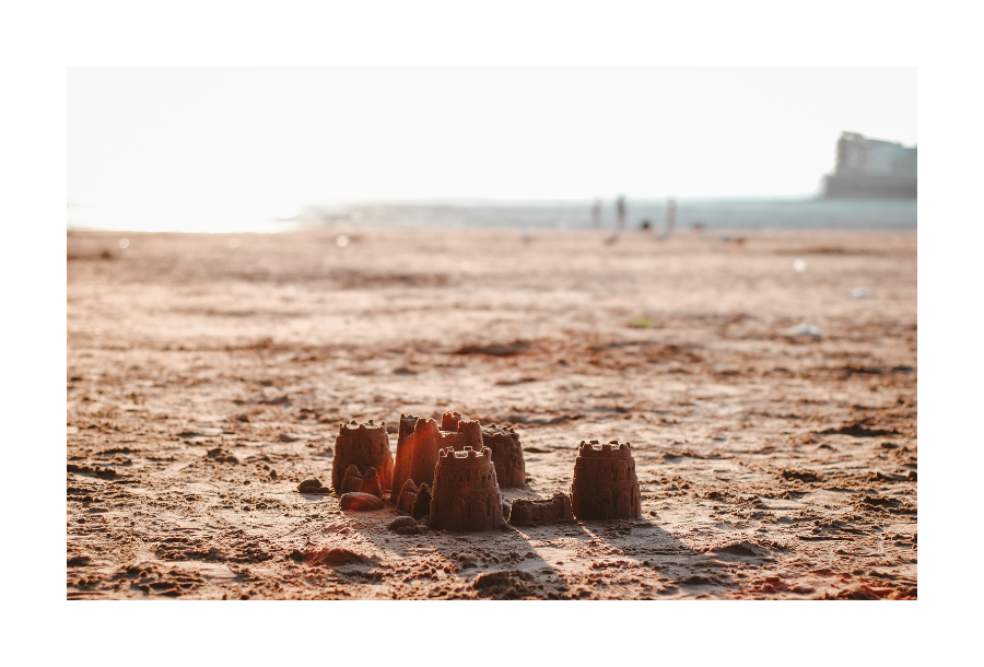 Pictured, a beautiful beach at dusk, muted pictures in the background of people by the water and in the foreground is a pile of sandcastles.