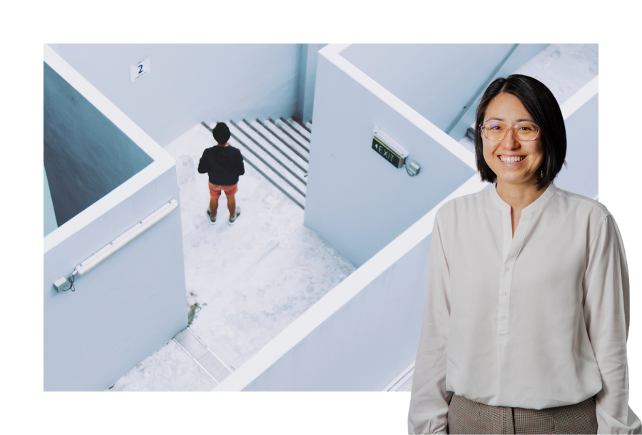 Pictured, Helen Yau our SMSF Manager and Financial Planner standing in front of a stock picture featuring white walls that represent a maze, with a person standing looking at a set of stairs next to an exit sign.