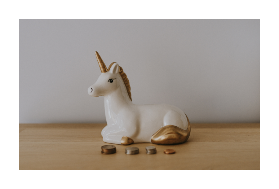 Pictured, sitting gently on a wooden bench top, a ceramic white and gold unicorn that is a money box. There are a couple of brass coloured coins sitting on the bench in front of the unicorn.