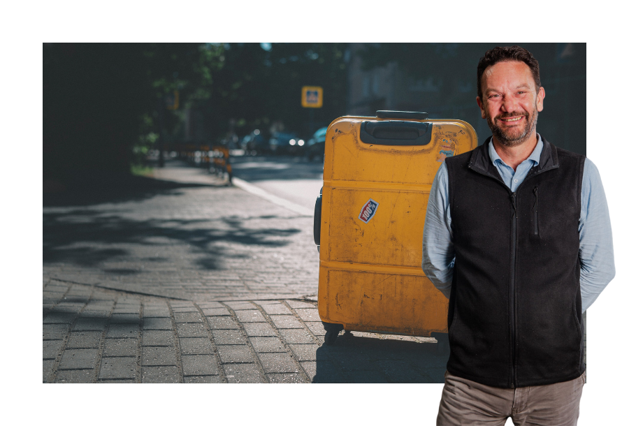 Pictured, Danny Grigg standing in front of a stock picture of a yellow suitcase sitting on the sidewalk.