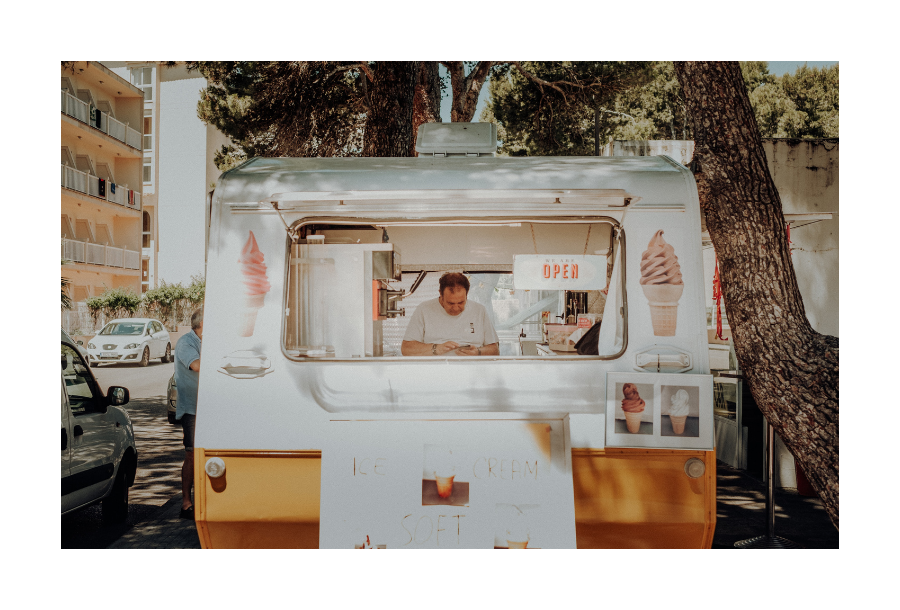 Pictured, a rear view of a white and orange caravan that has been transformed into a street cafe. You can see a person working inside on making a coffee on a busy street.