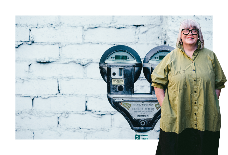 Pictured, an old school parking meter with a double head against a white painted brick wall. Standing in front of the picture is Anthea Taylor wearing a green top.