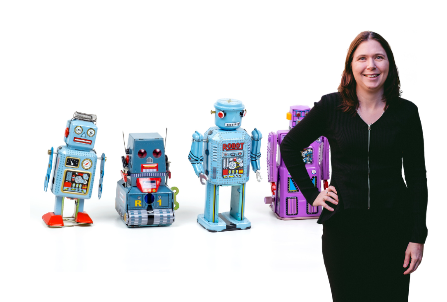 In front of a white background stand a number of different toy robots, with Karlene Wightman wearing a black dress at the front on the right hand side.