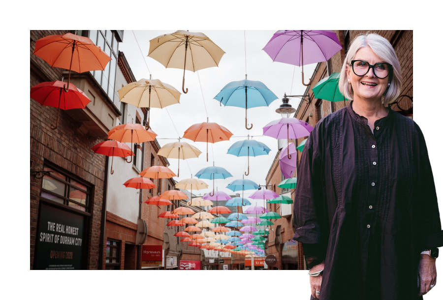 Pictured, Director Amanda Wilkens standing in front of a stock picture of beautiful bright coloured umbrellas being strung between two brick buildings.