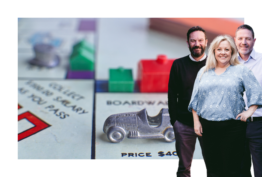 Pictured: Canny Accounting Managers, Danny, Michelle and Adam standing smiling in front of an image of a close up picture of a Monopoly game with a silver car on a square with a small green house and a larger red house.