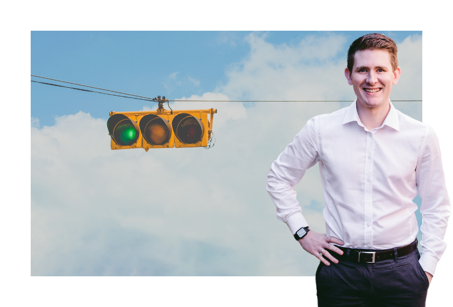 Pictured, an American style traffic light, with the green light flashing in front of the background of clouds. Standing in front is Chris Graham with one hand on his hip.