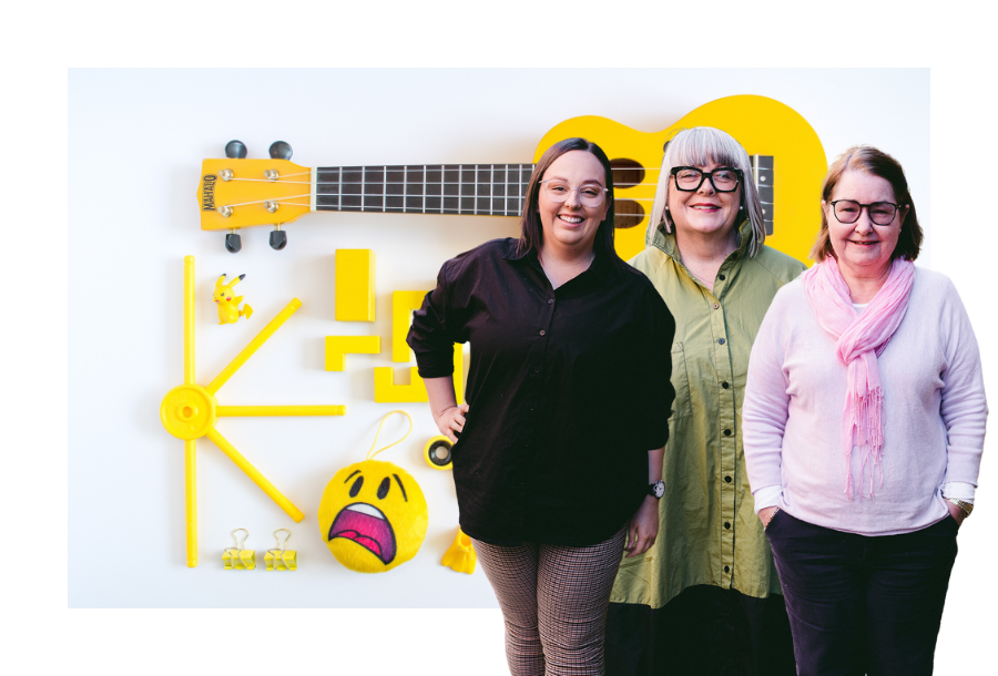 Mekayla Lamberty, Anthea Taylor and Christine Friel standing in front of a stock picture of bright yellow toys all organised neatly in a flat-lay.