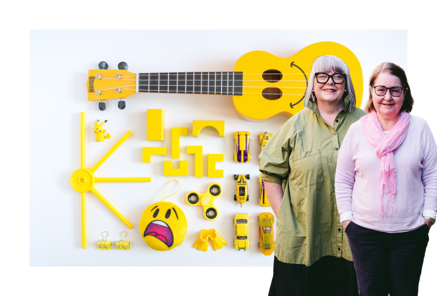 Pictured: Anthea Taylor and Christine Friel standing in front of a stock picture of bright yellow toys all organised neatly in a flat-lay.