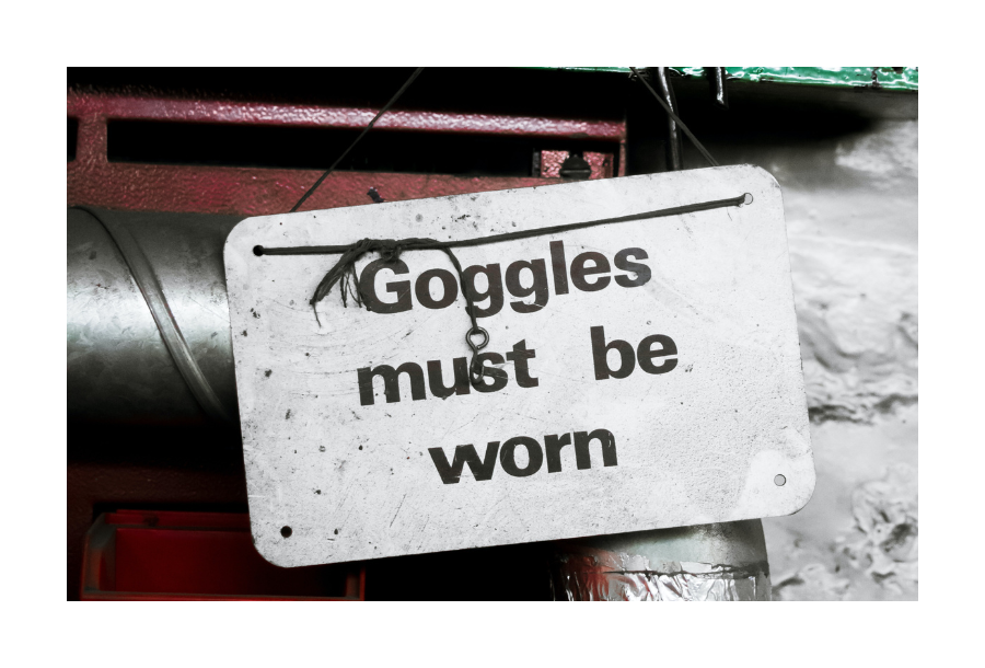 Pictured, a worn sign with the words that read "googles must be worm" - it is white in colour with black writing.