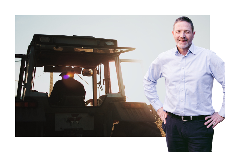 A picture of a tractor driving away with a man sitting in the driving seat at sunset. Standing with his hands on his hips, Adam Ramage wears a white shirt and black trousers.