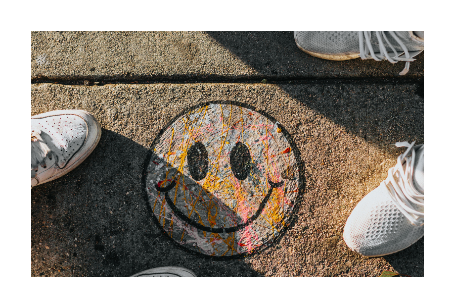 Pictured, a concrete path with two people standing opposite each other with white shoes. In between them, painted on the ground is a black outline of a smiley face.