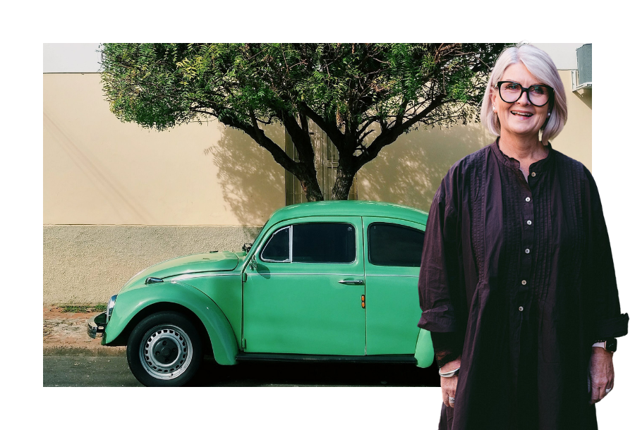 Pictured, Amanda Wilkens wearing a long sleeve black dress and thick framed black glasses. Standing in front of a picture of a green coloured Volkswagen Beeton parked on the street, next to a tree.