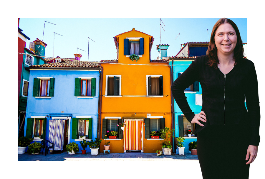Pictured Karlene Wightman with one hand on her hip wearing a black dress, standing in front of a stock picture of three houses, all bright colours of blue, orange and and green with a blue sunny sky.
