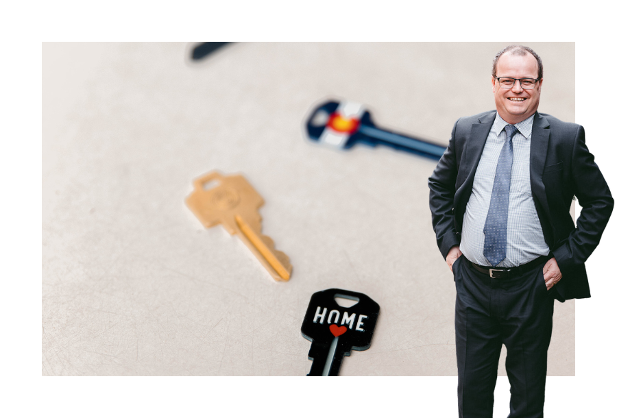 Pictured, Adam Wightman wearing a dark coloured suit with his hands in his pockets. Standing in front of a stock picture of flat layed house keys, with a black key and the words "home" with a red love heart.